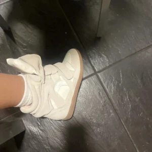 Vita sneakers med hög plös och kardborre - Säljer ett par vita sneakers med hög plös och kardborreband. Skorna har en chunky sula i beige och är tillverkade i syntetmaterial. Perfekta för dig som vill sticka ut med en unik och cool stil.