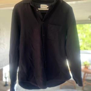 Svart skjorta från East West - Säljer en svart långärmad skjorta från East West. Skjortan har klassisk krage, bröstficka och knäppning framtill. Materialet känns lite grövre och strukturerat, perfekt för en stilren look. Storlek S