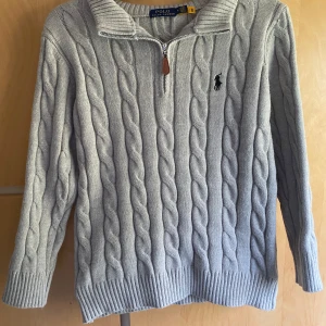 Ljusgrå kabelstickad tröja från Polo Ralph Lauren - Säljer en ljusgrå kabelstickad tröja från Polo Ralph Lauren med halv dragkedja och hög krage. Tröjan har ribbade muddar och det klassiska broderade logotypen på bröstet. Tillverkad i 100% bomull.