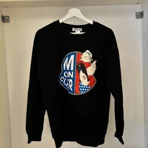 Svart Moncler tröja med Popeye-tryck - Cool svart tröja från Moncler med stort Popeye-tryck på bröstet och Moncler-logga på ärmen. Tröjan har rund hals och långa ärmar. Perfekt för dig som vill sticka ut med en unik design.