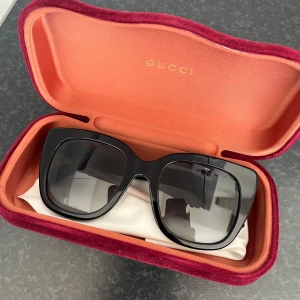 Svarta solglasögon från Gucci - Säljer ett par snygga svarta solglasögon från Gucci med bred båge och fyrkantig form. Glasögonen har diskreta gulddetaljer på skalmarna och kommer i ett lyxigt vinrött fodral med sammet.