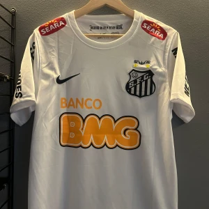 Vit Santos FC Neymar Jr fotbollströja - Säljer en vit Santos FC fotbollströja med Neymar Jr och nummer 11 på ryggen. Tröjan har svarta tryck, orange BMG-logga och klubbmärke på bröstet. Kortärmad modell i lätt material, perfekt för fotboll eller samling. Helt ny med tagg en replika 