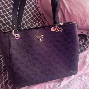 Svart axelväska från Guess - Säljer en svart axelväska från Guess med diskret logomönster och silvriga metalldetaljer. Väskan har dubbla handtag och dragkedja upptill. Perfekt för dig som vill ha en stilren och rymlig väska med klassisk design. En perfekt skick. Inga spår av användning. Ursprungligt pris cirka 120 €. Den är äkta.