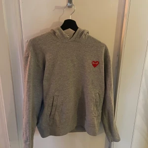 Grå hoodie från Comme des Garçons Play - Hej, jag säljer en grå hoodie från Comme des Garçons med det ikoniska röda hjärtat broderat på bröstet. Hoodien är väldigt bekväm och perfekt för en avslappnad stil. Dessutom är den i toppskick då den inte har nå defekter eller fläckar. Buda gärna! 