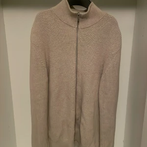 Beige stickad kofta med dragkedja - Säljer en beige stickad kofta med hög krage och dragkedja framtill. Tröjan har lång ärm och är ribbstickad vid ärmslut och nederkant. Perfekt för kyligare dagar och enkel att matcha med olika outfits. Priset kan diskuteras