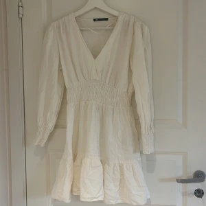 klänning i linne - Säljer en söt vit klänning från Zara med v-ringning och långa ärmar. Klänningen har smock i midjan och vid ärmslut samt volang nertill. Perfekt för vår och sommar, enkel att styla med sneakers eller sandaler. Den är i linne. Defekter förekommer