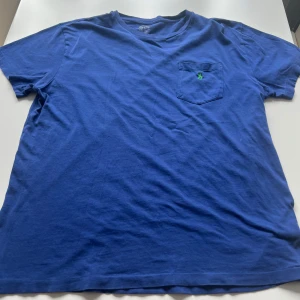 Blå t-shirt från Polo Ralph Lauren - Säljer en blå t-shirt från Polo Ralph Lauren med klassisk passform. T-shirten har en bröstficka med grön broderad logga och är tillverkad i mjuk bomull. Perfekt för en avslappnad stil.