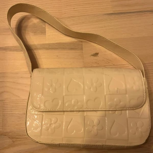 Beige axelväska med hjärtan och blommor - Säljer en beige axelväska med glansig finish och präglat mönster av hjärtan och blommor. Väskan har ett lock och bred axelrem, perfekt för att bära det viktigaste. Enkel och söt design som sticker ut.