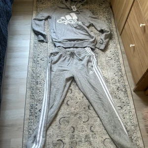Grå hoodie från Adidas och byxor - Säljer en grå hoodie från Adidas och byxor med  stor vit logga på bröstet med vita ränder längs sidorna. Tröjan har huva och långa ärmar, och är tillverkad i mjuk bomullsmix. Perfekt för en sportig och avslappnad stil.