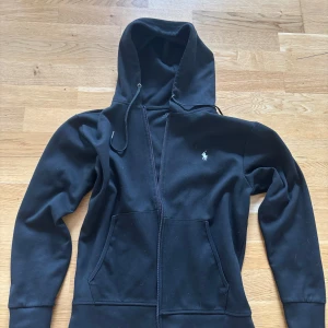 Svart hoodie från Polo Ralph Lauren - Säljer en svart hoodie från Polo Ralph Lauren med dragkedja och klassisk broderad logga på bröstet. Tröjan har huva med snörning och två fickor framtill. Perfekt för en avslappnad stil. Tröjan är i mycket bra skick.