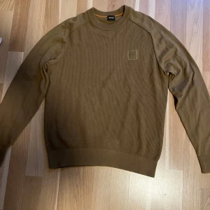 Brun stickad tröja från BOSS - Säljer en brun stickad tröja från BOSS med rund halsringning och diskret logotyp på bröstet. Tröjan har lång ärm och ribbade muddar vid ärmslut och nederkant. Perfekt för lager-på-lager och stilrena outfits. Obs endast anv 1-2 ggr så nyskick! Strl S 