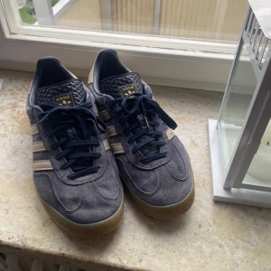 Adidas Gazelle sneakers i mörkblå mocka - Säljer ett par Adidas Gazelle sneakers i mörkblå mocka med klassiska vita ränder och gummisula. Skorna har snörning och gulddetaljer på sidan. Perfekta för dig som gillar retrostil och vill ha något bekvämt till vardags. Säljer dem endast för att dem är för små för mig. Hade aldrig nånsin sålt dem annars. Otroligt bra skicka som man ser, skorna går nästan inte heller att får tag i. 