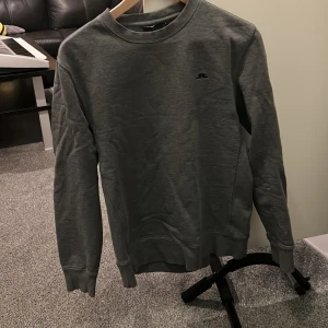 Mörkgrå sweatshirt från J.Lindeberg - En stilren mörkgrå sweatshirt från J.Lindeberg med rund halsringning och diskret logga på bröstet. Tröjan har långa ärmar och ribbade muddar vid ärmslut och nederkant. Perfekt för en avslappnad look.