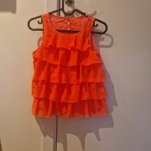 Orange volangtopp - Säljer en orange ärmlös topp med flera volanglager framtill. Toppen har rund halsringning och knäppning bak i nacken. Perfekt för sommarens varma dagar.