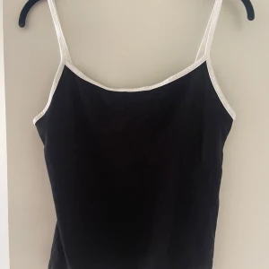 Svart linnetopp med vita band - Säljer en svart linne med tunna vita axelband och vit kant runt halsen. Perfekt basplagg till sommaren och enkel att matcha med olika outfits. Endast använd 1 gång 