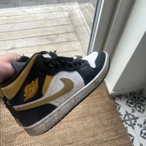 Nike Air Jordans svart, vit och guld - Säljer ett par Nike Air Jordans i svart, vit och guld med klassisk vinge-logga på sidan. Skorna har snörning, platt sula och rund tå. Perfekta för dig som gillar streetwear och vill sticka ut med snygga färgdetaljer.