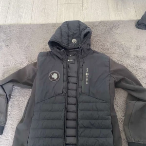 Svart hybridjacka med huva från Sail Racing - Säljer en svart Sail Racing Antarctica Hybrid Hood med quiltade partier och huva. Jackan har dragkedja framtill, två sidofickor och Sail Racing-logga på bröstet. Perfekt för kyliga och medelvarma dagar och snygg look.