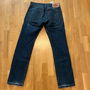 Levi's 501 jeans med rå kant - Mörkblå Levi's 501 jeans med raka ben och klassisk femficksdesign. Jeansen har rå, fransad kant nedtill och tydliga kontrastsömmar. Ikonisk läderpatch bak i midjan och röd Levi's-tagg på bakfickan. Midjemått W32 och längd L32.