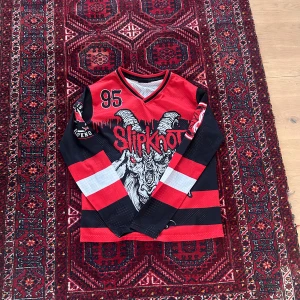 Slipknot långärmad tröja med tryck - Säljer en röd och svart långärmad tröja från Slipknot med stort tryck av getskalle och bandets logga framtill. Tröjan har V-ringning, svarta och röda ränder samt vita detaljer på ärmarna. Perfekt för dig som gillar bandmerch!