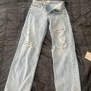 Ljusblå jeans med raka ben och hål - Säljer ett par ljusblå jeans med raka ben och slitningar på båda benen. Jeansen har klassisk femficksdesign, knappgylf och hög midja. Perfekta för en avslappnad och trendig look.