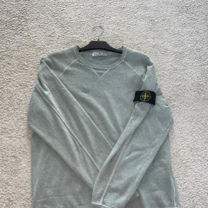 Ljusgrå sweatshirt från Stone Island - Säljer en stilren ljusgrå sweatshirt från Stone Island med klassisk patch på vänster ärm. Tröjan har rund halsringning och långa ärmar. Perfekt för en avslappnad och trendig look.