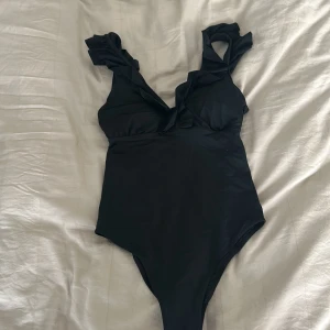 Svart baddräkt med volangdetaljer - Aldrig använd som xs eller S.Säljer en svart baddräkt med breda axelband och volangdetaljer längs banden och urringningen. Baddräkten har en liten öppning framtill och är tillverkad i ett stretchigt syntetmaterial. Perfekt för stranden eller poolen!