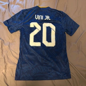 Real Madrid bortatröja Vini Jr. 20 - Mörkblå Real Madrid bortatröja med gula detaljer och tryck Vini Jr. 20 på ryggen. Adidas-logga och klubbmärke på bröstet samt sponsortryck på framsidan. Kortärmad modell i lätt och ventilerande material.