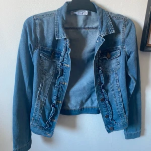 Jeansjacka med volang från Black Story - Säljer en snygg blå jeansjacka från Black Story i storlek XS. Jackan har långa ärmar, klassisk krage, knappar framtill och volangdetaljer längs knappraden. Perfekt för att lyfta din outfit med en cool touch.Frakt tillkommer