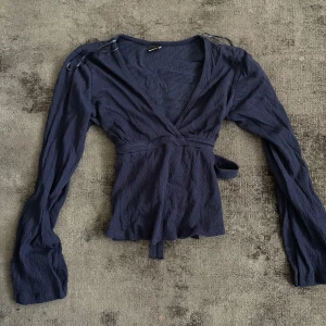 Mörkblå omlottopp med knytband - Säljer en mörkblå omlottopp med långa ärmar och knytband i midjan. Toppen har v-ringning och är tillverkad i ett lätt, strukturerat tyg. Perfekt för dig som gillar en feminin och trendig stil. 