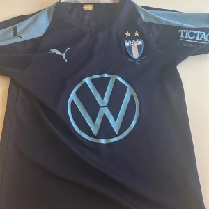 Malmö FF fotbollströja Puma - Säljer en officiell Malmö FF fotbollströja från Puma i mörkblått med ljusblå detaljer. Tröjan har klubbmärke, två stjärnor, Volkswagen-logga på bröstet och Tillmobil-tryck på ryggen. Kortärmad modell med V-ringning och broderad svensk flagga i nacken. Tröjan är i storlek 152/ 11–12 år