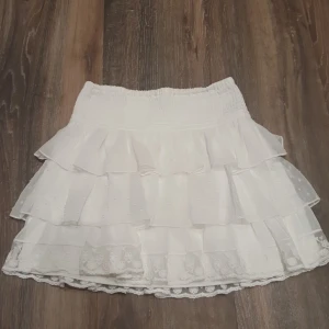 Vit volangkjol från Lindex - Supersöt vit kjol från Lindex med flera lager volanger och spetsdetaljer längst ner. Kjolen har bred resår i midjan och är kort i modellen. Perfekt för dig som gillar romantisk och lekfull stil.