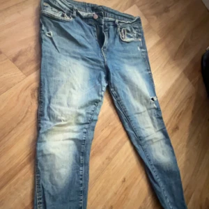 Blå  skinny slitna jeans med hål - Säljer ett par blå jeans med slitningar och hål på benen. Jeansen har klassisk femficksdesign, dragkedja och knapp i midjan. De har en något smalare passform och är tillverkade i denim. Perfekta för en avslappnad stil.