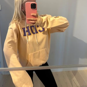 Gul hoodie från Champion med lila tryck - Säljer en gul hoodie från Champion med lila text framtill. Tröjan har magficka och huva med dragsko. Perfekt för en avslappnad stil och riktigt skön att ha på sig.