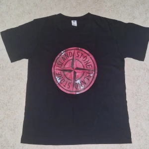Svart t-shirt med röd Stone Island-logga - Säljer en svart t-shirt från Stone Island med stor röd logga på bröstet. T-shirten har rund hals och korta ärmar. Tillverkad i bomull och passar perfekt för en avslappnad stil.