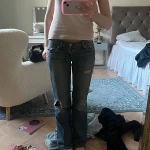 Blå slitna bootcut jeans - Säljer ett par blå bootcut jeans från Levi’s med slitningar och hål på knä och lår. Jeansen har låg midja, klassiska fickor och dragkedja. 
