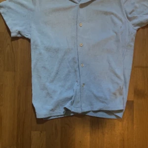 Ljusblå frottéskjorta med knappar - Säljer en ljusblå skjorta i mjuk frotté med korta ärmar och klassisk krage. Skjortan har knäppning framtill med fyra knappar och avslappnad passform. Perfekt för varma dagar eller som ett chill plagg över t-shirt.