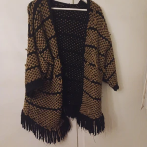 Stickad cardigan jacka med fransar - Säljer en stickad cardigan jacka i svart och beige med coolt rutigt mönster och långa fransar nedtill. Jackan har en loose passform och trekvartsärm, perfekt att slänga på sig för en avslappnad vibe.