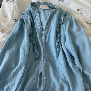 Blå volangskjorta från Neo Noir - Supersöt ljusblå skjorta från Neo Noir med volangdetaljer längs axlar och bröst. Skjortan har knappar framtill, lång ärm och en lätt oversized passform. 