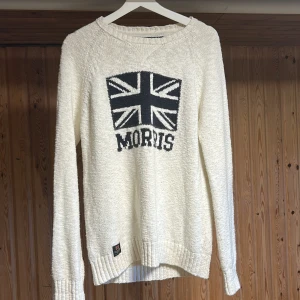 Vit stickad tröja från Morris - Vit stickad tröja från Morris med Union Jack-flagga och texten 'Morris' på bröstet. Tröjan har rund hals, ribbade muddar och är långärmad. Perfekt för dig som gillar klassisk brittisk stil och vill ha något som sticker ut.
