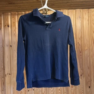 Mörkblå pikétröja från Ralph Lauren - Klassisk mörkblå långärmad pikétröja från Polo Ralph Lauren med röd broderad logga på bröstet. Tröjan har krage, knäppning vid halsen och är tillverkad i mjuk bomull. Perfekt för en stilren och avslappnad look.