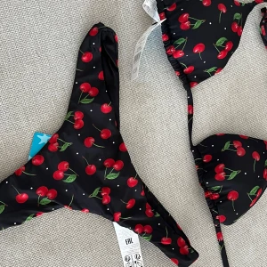 Svart bikini med körsbär från Tezenis - Supersöt svart bikini från Tezenis med körsbärsmönster och små vita prickar. Klassisk trekantsmodell upptill med knytband och matchande trosa med hög benskärning. Strl xs/s