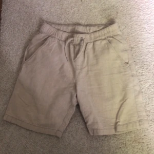 Beiga linneshorts från Kappahl - Snygga beiga shorts från Kappahl i storlek 152. De är gjorda i linne, har elastisk midja med snörning och två sidfickor fram samt två bakfickor. Perfekta för varma dagar när du vill ha en chill och stilren look.