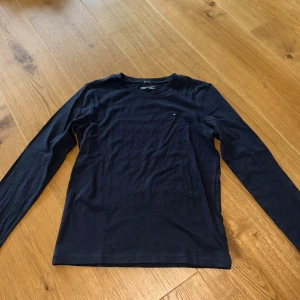2-pack Tommy Hilfiger långärmade tröjor - Två stilrena långärmade tröjor från Tommy Hilfiger i storlek 152. En marinblå och en vit, båda i mjuk ekologisk bomull. Klassisk passform med rund hals och diskret logga på bröstet. Perfekta för en clean och enkel look.