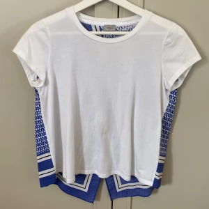 Vit och blå t-shirt från Marella Sport - Snygg t-shirt från Marella Sport med vit framsida och blåvit mönstrad baksida. Kortärmad modell i bomull med coolt grafiskt mönster och randiga detaljer bak. Unik omlott-effekt baktill som ger extra edge till looken. Liten fläck på framsidan, se bild 5.