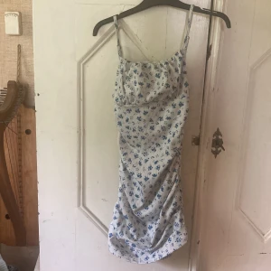 Blommig kortklänning från SHEIN -  Kortklänning från SHEIN med smala axelband och blått blommönster på vit botten. Klänningen är figurnära och har rynkade detaljer längs sidorna. Tillverkad i stretchig polyesterblandning.