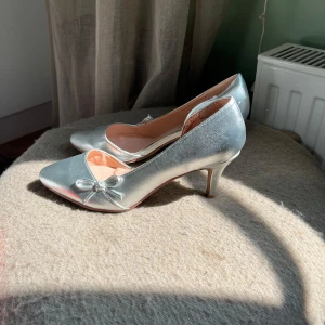 Silvriga pumps med rosett från BODYFLIRT - Snygga pumps i metallic-silver från BODYFLIRT med spetsig tå och liten rosett framtill. Skorna har låg klack & är bara använda en gång .  Perfekta för dig som vill sticka ut med stilrena detaljer.