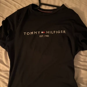 Svart t-shirt Tommy Hilfiger - Svart t-shirt från Tommy Hilfiger med klassisk logga och texten 'EST. 1985' broderad på bröstet. Enkel och stilren design med rund hals och korta ärmar. Perfekt för en clean och avslappnad look.