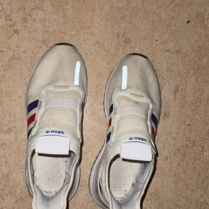 Vita Adidas sneakers med blå och röda ränder - Snygga vita Adidas sneakers med klassiska blå och röda ränder på sidorna. Skorna har en stickad ovandel i syntetmaterial och elastiska band istället för traditionell snörning. Perfekta för en sportig och avslappnad stil.