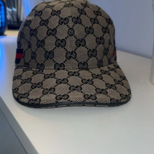 Gucci keps med GG-mönster och röd/blå detalj - Exklusiv keps från Gucci i beige och svart med ikoniskt GG-mönster över hela kepsen. Snygg röd och marinblå rand på sidan. Justerbar rem baktill och insida i mörkblått tyg. Perfekt för dig som vill sticka ut med lyxig streetstyle. Storlek M men ändå justerbar pris 900 pris kan diskuteras 