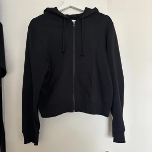 Svart hoodie med dragkedja och huva - Svart hoodie med dragkedja framtill, huva med snörning och två stora fickor. Klassisk modell med ribbade muddar och lång ärm. Perfekt för chill dagar eller när du vill ha en enkel och stilren look.